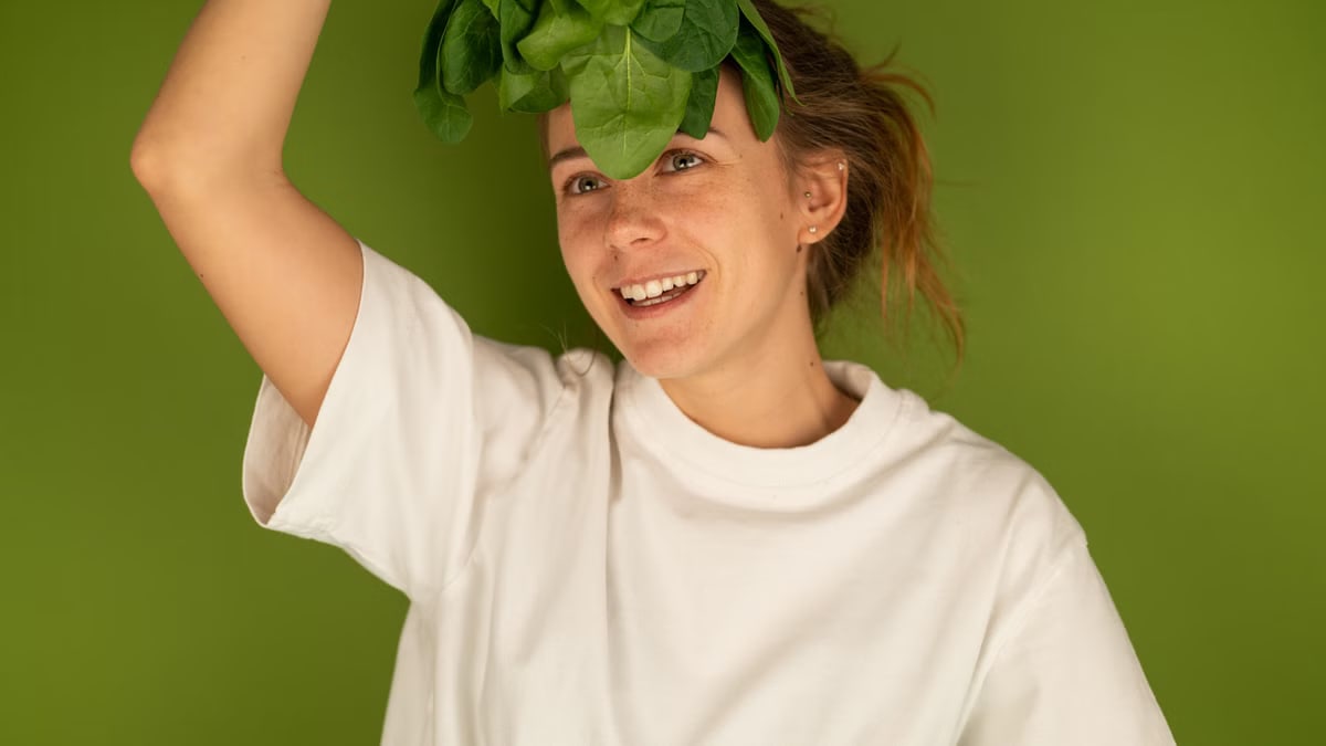 Cheerful adult female in white t shirt with fresh spinach foliage against forehead looking up